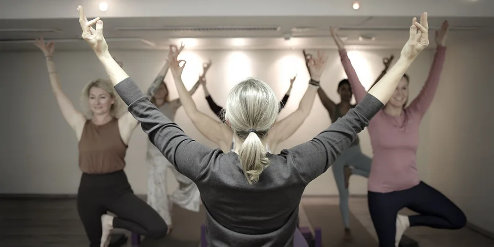 Ohjaaja etualalla selin kameraan ja ryhmä aikuisia tekemässä joogaliikettä (puu-asento) valoisassa joogasalissa, kädet ylhäällä tasapainoasennossa.