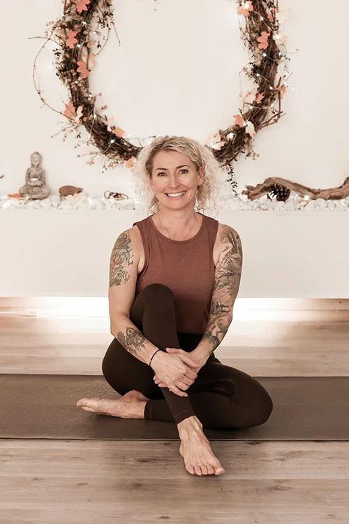 Hymyilevä nainen istuu risti-istunnassa joogamatolla rauhallisessa sisätilassa. Hänellä on lyhyet vaaleat hiukset, hihaton paita ja tatuoinnit käsivarsissa. Taustalla on koristeellinen kranssi ja pieniä sisustuselementtejä.