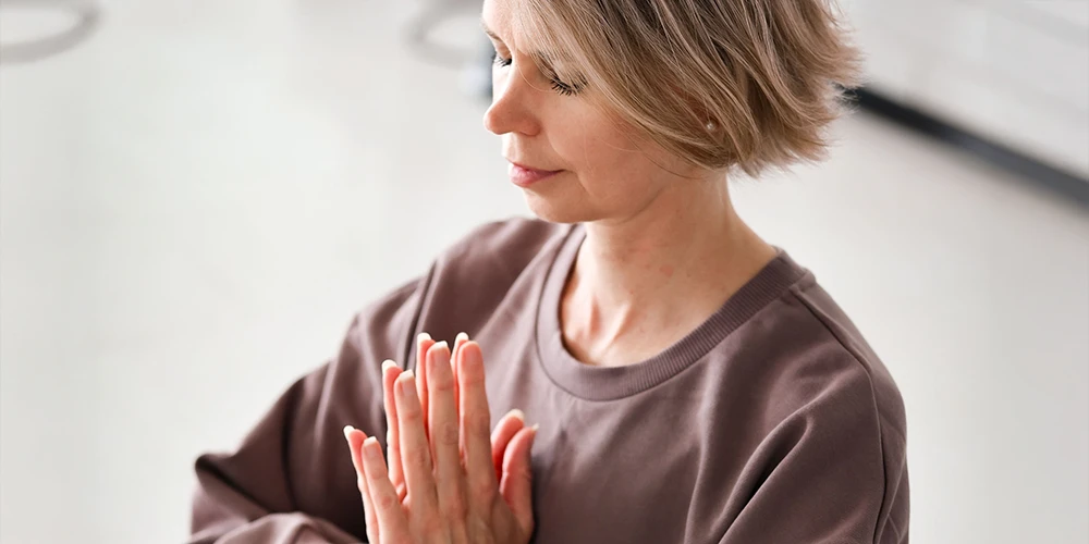 Lyhythiuksinen henkilö istuu joogamatolla sisätilassa, kädet rukousasennossa rinnan edessä ja silmät suljettuina rauhallisessa joogameditaatiohetkessä.